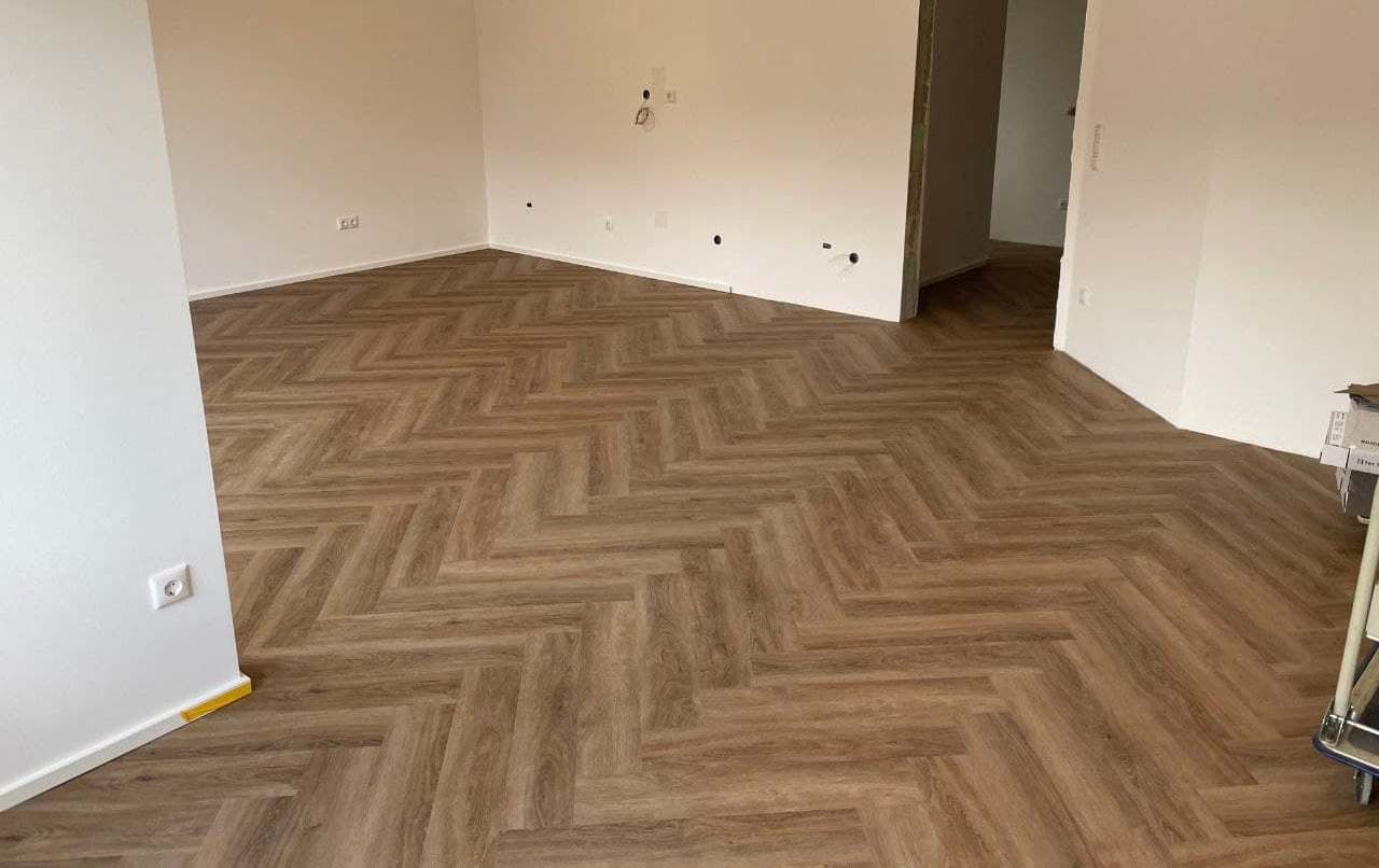 Fertig verlegter Designboden im Fischgrätmuster in einem hellen, leeren Wohnraum, die Fläche läuft bis in den angrenzenden Flur durch