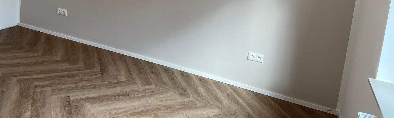 Nahaufnahme des Wandanschlusses: der Designboden im Fischgrätmuster endet mit einer schmalen Randfuge an der weißen Sockelleiste