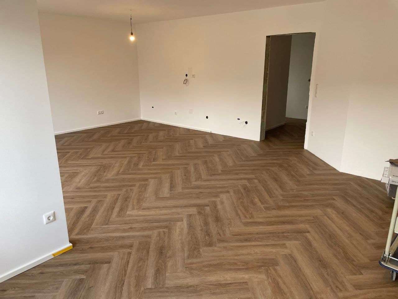 Leerer heller Wohnraum mit frisch verlegtem Designboden im Fischgrätmuster, die Fläche reicht bis in den angrenzenden Raum