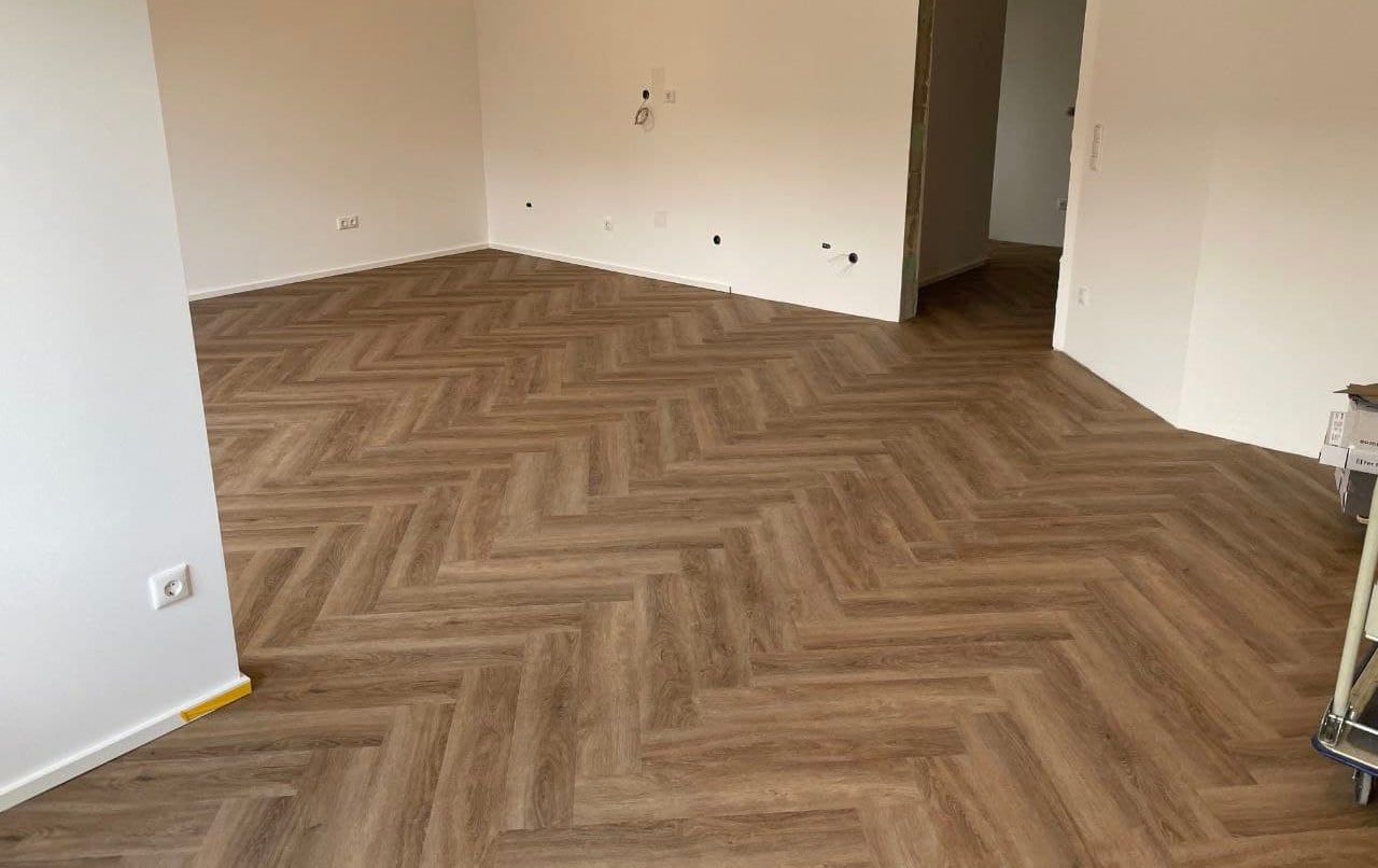 Fertig verlegter Designboden im Fischgrätmuster in einem hellen, leeren Wohnraum, die Fläche läuft bis in den angrenzenden Flur durch